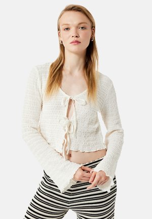 Ung kvinde iført en tekstureret hvid crop top med lange ærmer og bindedetaljer samt sort-hvide stribede bukser, stående mod en ensfarvet baggrund.