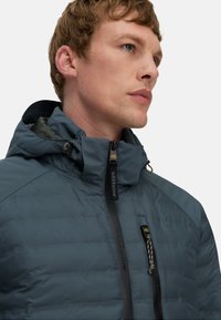 Padded jacket w ciemnym morskim kolorze, z wysokim kołnierzem, zamek błyskawiczny z przodu i regulowaną kapturem. Widoczne są charakterystyczne szwy i akcenty z logo.