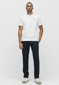 Hvid bomulds T-shirt med et lille rødt logo og korte ærmer, parret med mørkeblå denim jeans og lyse sneakers.