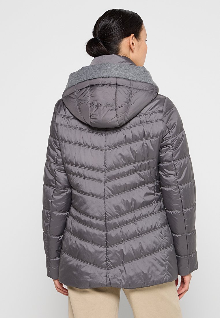 Kobieta w szarej pikowanej kurtce z kapturem, plecami do nas, w beżowych spodniach i z włosami spiętymi w niskiego koka.