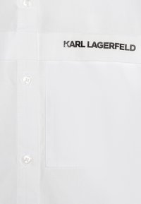 Bela srajca z gumbki iz gladkega bombaža z rahlim leskom. Ima žep na levi strani prsnega dela in črno izvezeno znamenje "KARL LAGERFELD".