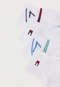 Τέσσερα ζευγάρια λευκές κάλτσες αστραγάλου με διαφορετικά χρωματιστά λογότυπα σε ρίγες και κεντημένα σχέδια στις μανσέτες, τοποθετημένα διαγώνια.