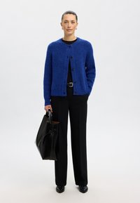 Kardigan w kolorze królewskiego błękitu o fakturze, zapinany na czarne guziki, stylizowany na czarną bluzkę, zestawiony z czarnymi spodniami z wysokim stanem oraz czarną torbą typu tote.