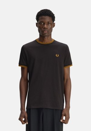Hombre con cabello corto y rizado que lleva una camiseta negra con ribete amarillo en el cuello y las mangas, de pie frente a un fondo liso.