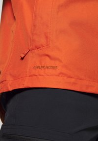 Gros plan d'une veste de sport orange avec le texte "CEPLEX ACTIVE" au-dessus d'un pantalon noir avec une poche zippée visible.