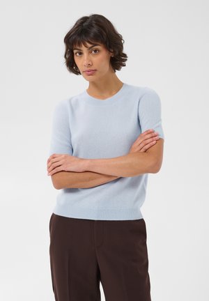 Femme aux cheveux courts et foncés portant un pull bleu clair à manches courtes et un pantalon marron foncé, debout les bras croisés devant un fond uni.
