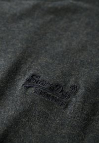 Tejido gris oscuro con un tejido texturizado. Presenta un logo bordado en azul marino, ubicado en la esquina inferior. Diseño suave y casual.