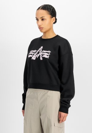 Mujer con el cabello trenzado que lleva una sudadera negra con un diseño floral rosa y pantalones cargo beige, de pie frente a un fondo blanco.