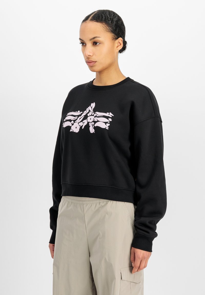 Femme aux cheveux tressés portant un sweat-shirt noir avec un motif floral rose et un pantalon cargo beige, debout devant un fond blanc.