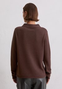 Brązowy sweter z dzianiny z kołnierzem, o luźnym kroju i ściągaczach u rękawów. Tekstura jest miękka, a materiał wydaje się lekki.