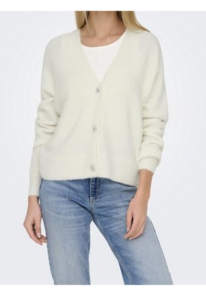 Vrouw draagt een witte V-hals cardigan met decoratieve knopen over een witte top, gecombineerd met lichtblauwe spijkerbroeken, tegen een effen achtergrond.