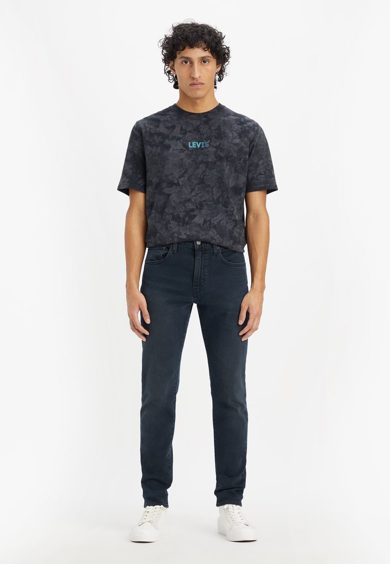 T-shirt noir à motif camouflage avec le logo Levi's en teal, associé à un jean foncé et des baskets blanches. Manches courtes et coupe décontractée.