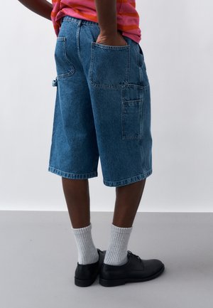 Άτομο που φοράει μπλε τζιν cargo σορτς, με τα χέρια στις πίσω τσέπες, λευκές κάλτσες και μαύρα επίσημα παπούτσια, στέκεται μπροστά σε απλό φόντο.