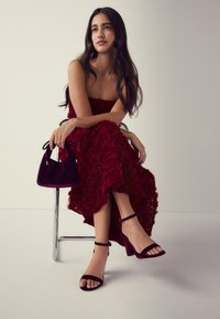 Burgundské květinové šaty s vrstvenou růžovou texturou, doplněné o ladící semišové podpatky a kabelku. Modelka sedí na chromové židli před světlým pozadím.