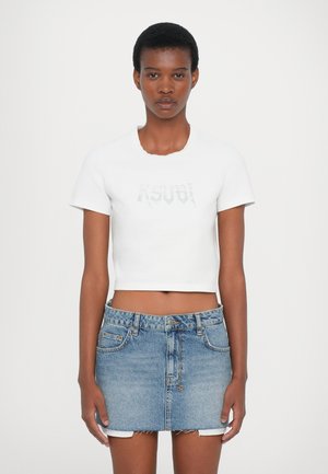 Camiseta blanca corta con cuello redondo y mangas cortas, con un logo plateado en el frente. Combinada con una minifalda de mezclilla azul.