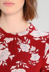 Roter strukturierter Stoff mit blauen Blumenmuster, mit rundem Ausschnitt und kurzen Ärmeln, das einen Teil des Oberkörpers eines Modells zeigt.