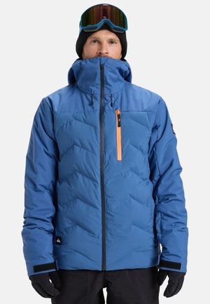 Homme portant une veste d'hiver bleue avec une fermeture éclair orange sur la poitrine, des gants noirs, un bonnet noir, et des lunettes de ski sur le front, faisant face à l'avant.