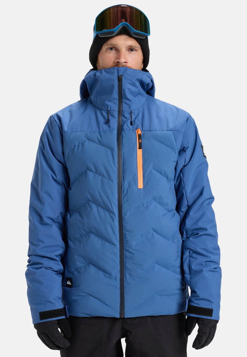 Mann in blauer Winterjacke mit orangefarbenem Reißverschluss auf der Brust, schwarzen Handschuhen, schwarzer Mütze und Skibrille auf der Stirn, der nach vorne schaut.