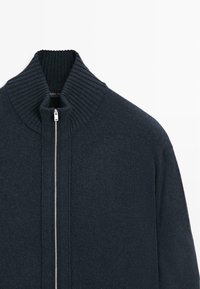 Maglione zip-up in maglione blu scuro con colletto alto, caratterizzato da una texture a coste e una zip in metallo argentato. Design semplice e moderno senza motivi aggiuntivi.