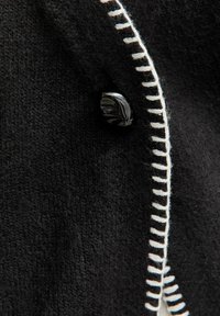 Czarna wełniana odzież z szerokim kremowym szwem i błyszczącym czarnym guzikiem w pobliżu krawędzi. Teksturowana tkanina dodaje wizualnego zainteresowania.