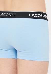Svetlo modri bokseric z črnim pasom, ki nosi belo blagovno znamko Lacoste, oblečen na spodnjem delu telesa in zgornjih stegnih osebe.