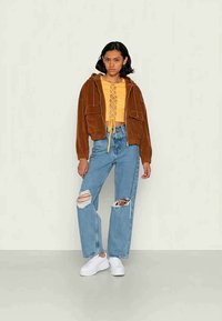 Brown corduroy cropped jacket, yellow lace-up top, light blue ripped jeans, white sneakers, standing against a light background.