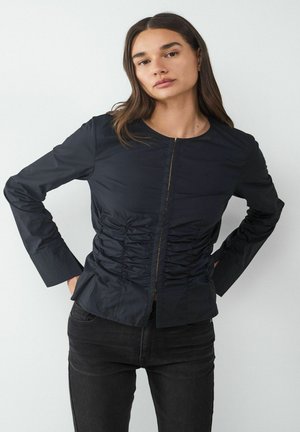 Žena v čiernej nariasenej blúzke s dlhým rukávom a čiernych džínsoch stojaca s rukami v bokoch na bielom pozadí.