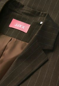 Rjava jopica v herringbone vzorcu z ozkimi vertikalnimi črtami. Ima rožnato oznako z napisom "JJXX" in gladko, svetlo rjavo notranjost.