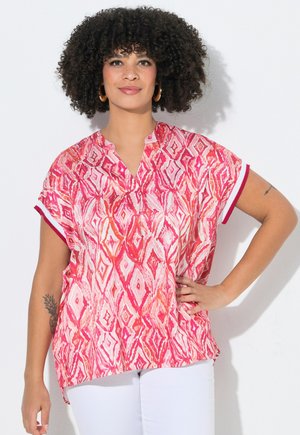 Kvinde med krøllet hår iført guldringe-øreringe, en pink mønstret kortærmet bluse og hvide bukser, stående med hånden på hoften.