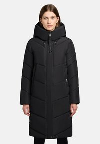 Frau, die einen langen schwarzen gepolsterten Wintermantel mit hohem Kragen und Kapuze trägt, steht vor einem schlichten hellen Hintergrund.