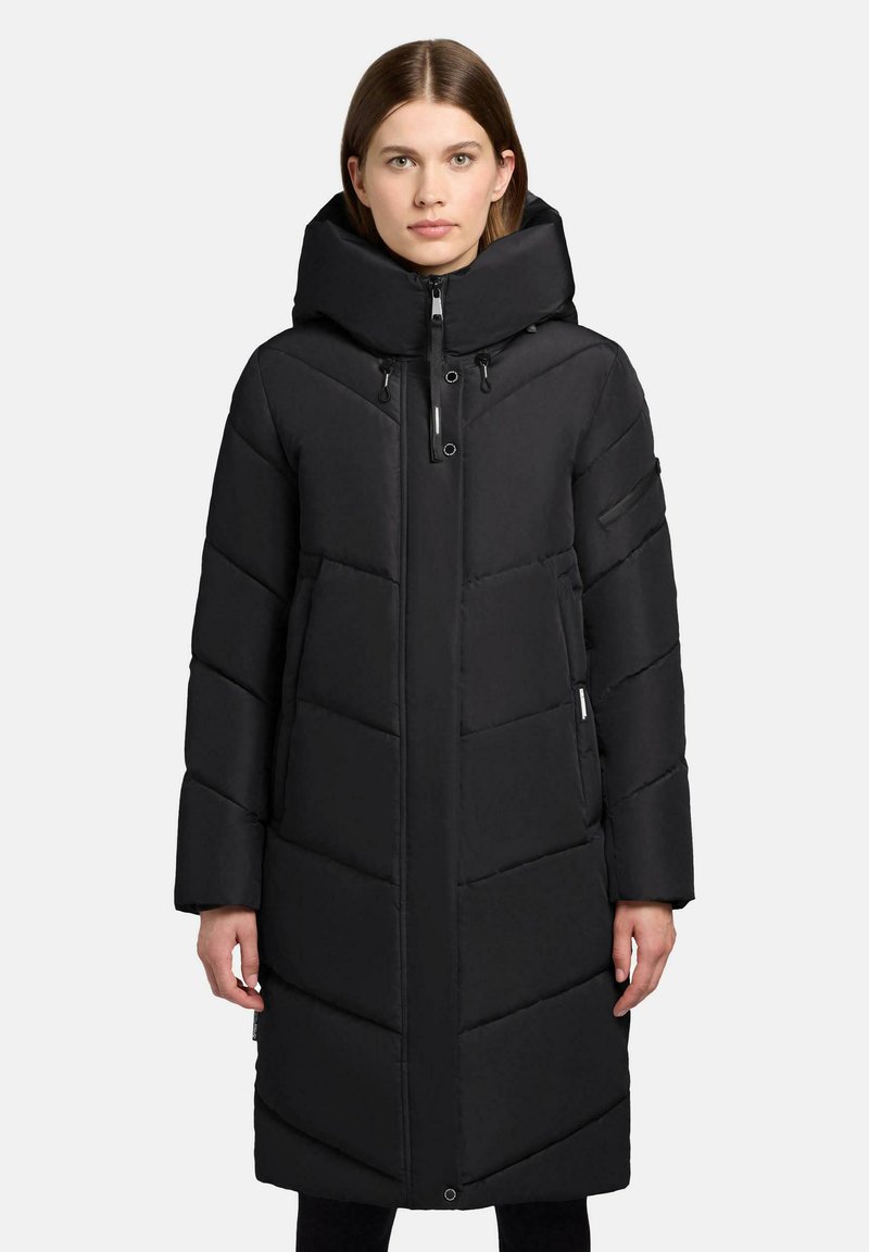 Frau, die einen langen schwarzen gepolsterten Wintermantel mit hohem Kragen und Kapuze trägt, steht vor einem schlichten hellen Hintergrund.