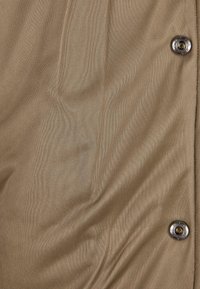 Lysebrunt stof med en glat tekstur, der har to metalliske knapper og diskrete folder som designdetalje.