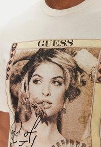 Camiseta de algodón blanca con un estampado gráfico del retrato de una mujer con acentos florales y el texto "GUESS," bordeado por patrones decorativos.