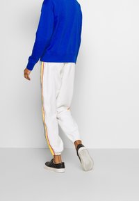 Vita sweatpants med flerfärgade ränder (röd, gul, blå) längs sidorna, tillsammans med en blå sweatshirt och svarta sneakers.