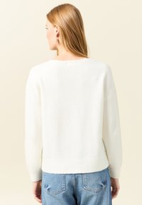 Witte, zachtgebreide trui met een relaxte pasvorm en ronde halslijn. Heeft lange mouwen en een geribde zoom, gedragen met blauwe jeans.