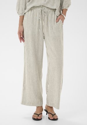 Frau trägt cremefarbene und schwarz vertikal gestreifte, weit geschnittene Hosen mit Kordelzug in der Taille, kombiniert mit schwarzen Sandalen mit Absatz und silbernem Armband.