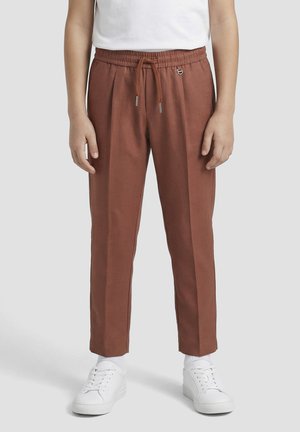 Niño con pantalones marrones estrechos con cintura elástica y cordón, combinados con zapatillas blancas y calcetines blancos, de pie sobre un fondo liso.