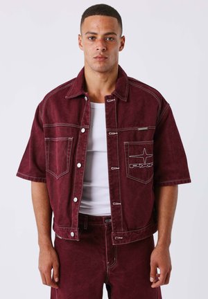 Man draagt een bordeauxrode korte mouw denim jas met witte stiksels over een wit hemd, gecombineerd met bijpassende bordeauxrode broek.