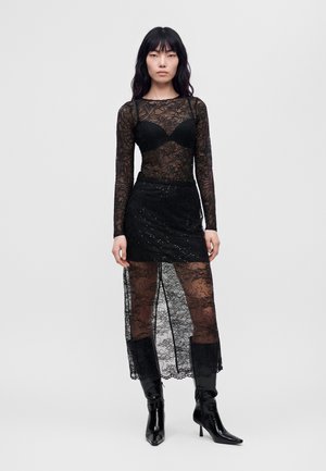 Un mannequin pose avec un haut noir en dentelle, une minijupe ornée, une superposition en dentelle transparente et des collants, ainsi que des bottes brillantes montant jusqu’aux genoux, sur un fond blanc uni.