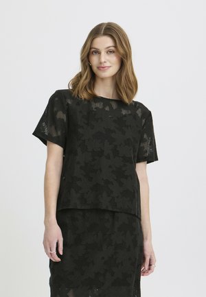 Femme aux cheveux châtain clair mi-longs portant un haut noir en dentelle florale à manches courtes et une jupe assortie, se tenant devant un fond uni.