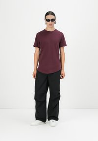 Camiseta marrón con mangas cortas y dobladillo redondeado, combinada con pantalones negros holgados que presentan un acabado texturizado y zapatillas blancas.