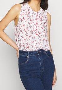 Blusa sin mangas en tejido ligero, blanca, con estampado floral en rojo y azul. Presenta un escote festoneado y un corte holgado, combinada con jeans azules.