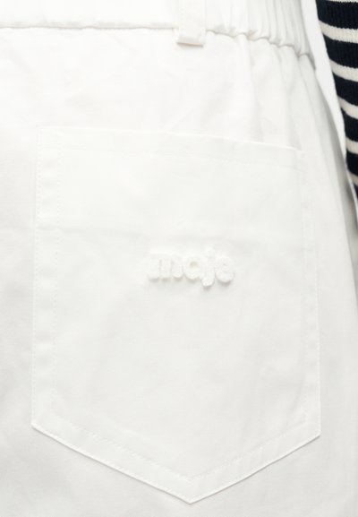 Poche en tissu blanc sur un vêtement avec une taille élastique, brodée du mot "maje" en fil blanc assorti.