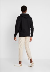Sudadera negra con cordón y bolsillo tipo canguro, combinada con pantalones chinos beige claros y zapatillas blancas con detalles en beige. Fondo blanco.