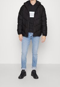 Giacca nera imbottita con cappuccio, dotata di chiusura zip e tasche laterali. Indossata sopra un polo nera e jeans azzurri a sigaretta con risvolti.