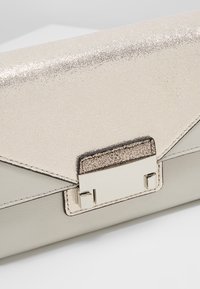 Kabelka přes rameno s metalickým třpytivým povrchem, béžová kůže, trojúhelníkový klapka a stříbrné kování. Čisté linie a minimalistický design.