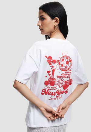 Hvid oversized t-shirt med et rødt grafisk print af et cocktailglas, kirsebær, terninger og en discokugle. Teksten lyder "New York Lovers Club."
