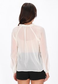 Blusa blanca transparente con mangas largas, textura suave, cuello con volantes y ajuste holgado, combinada con pantalones cortos negros.
