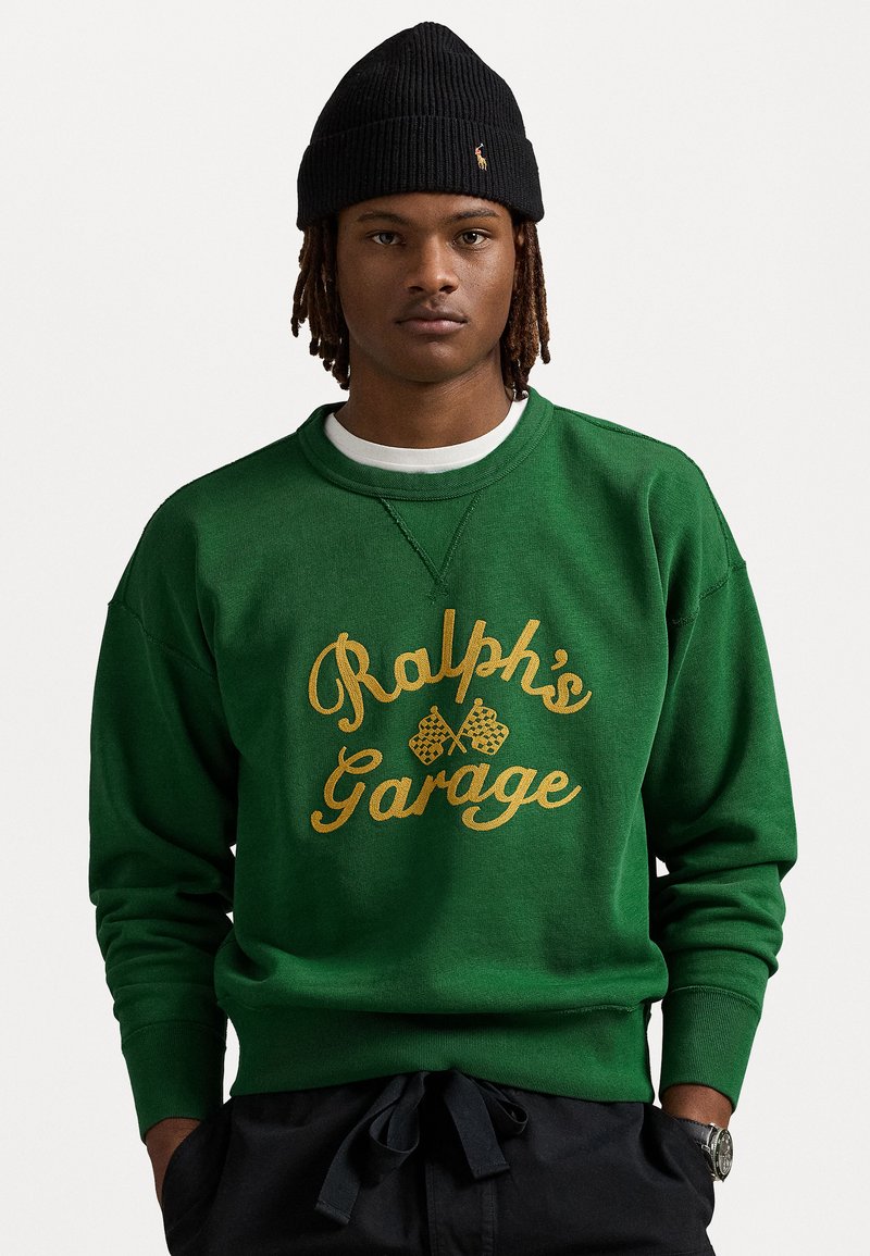 Green sweatshirt with gold "Ralph's Garage" print and racing flags; features ribbed neckline and cuffs, soft texture, worn over a white shirt.