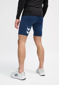 Man iklädd marinblåa sportshorts med vita chevron-logotyper, svart långärmad tröja och vita löparskor, stående på en vit bakgrund.